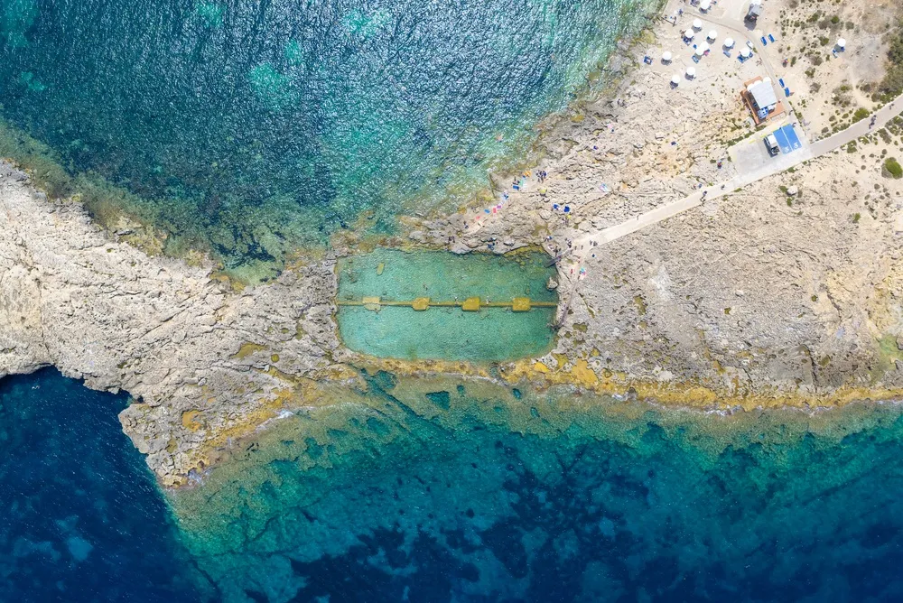 La piscina naturale di roccia a Qawra Point dall’alto, con bagnanti e acqua turchese